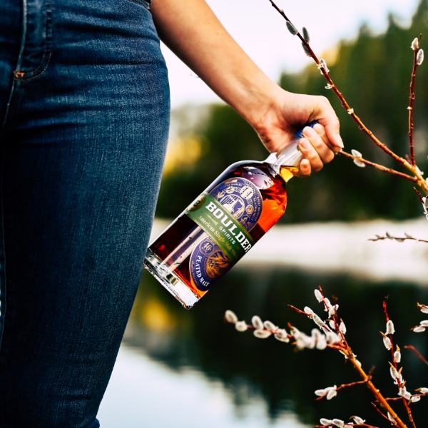 Woman in jeans holding Boulder whiskey bottle beside a brown plant with lake in the background