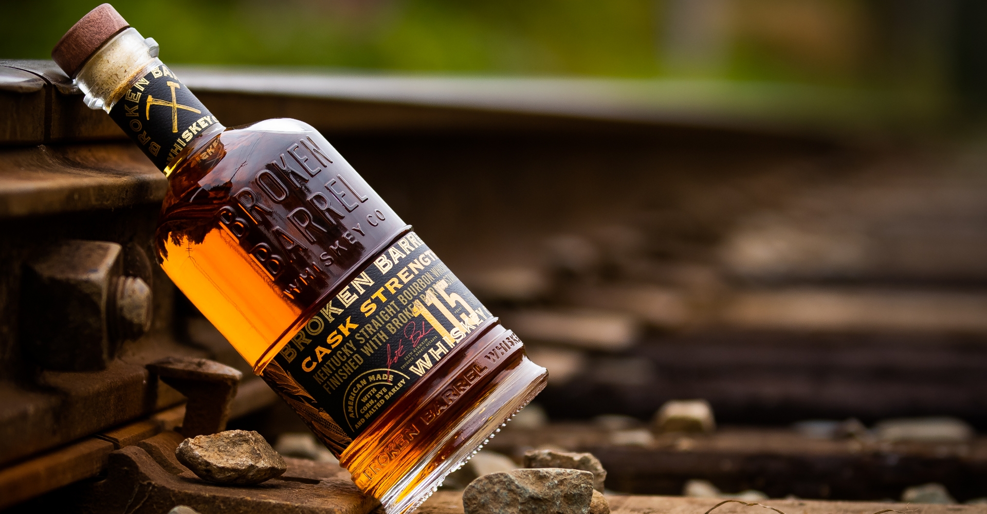 Broken Barrel Whiskey bottle leaning against rusty train tracks with slight green foliage in background.