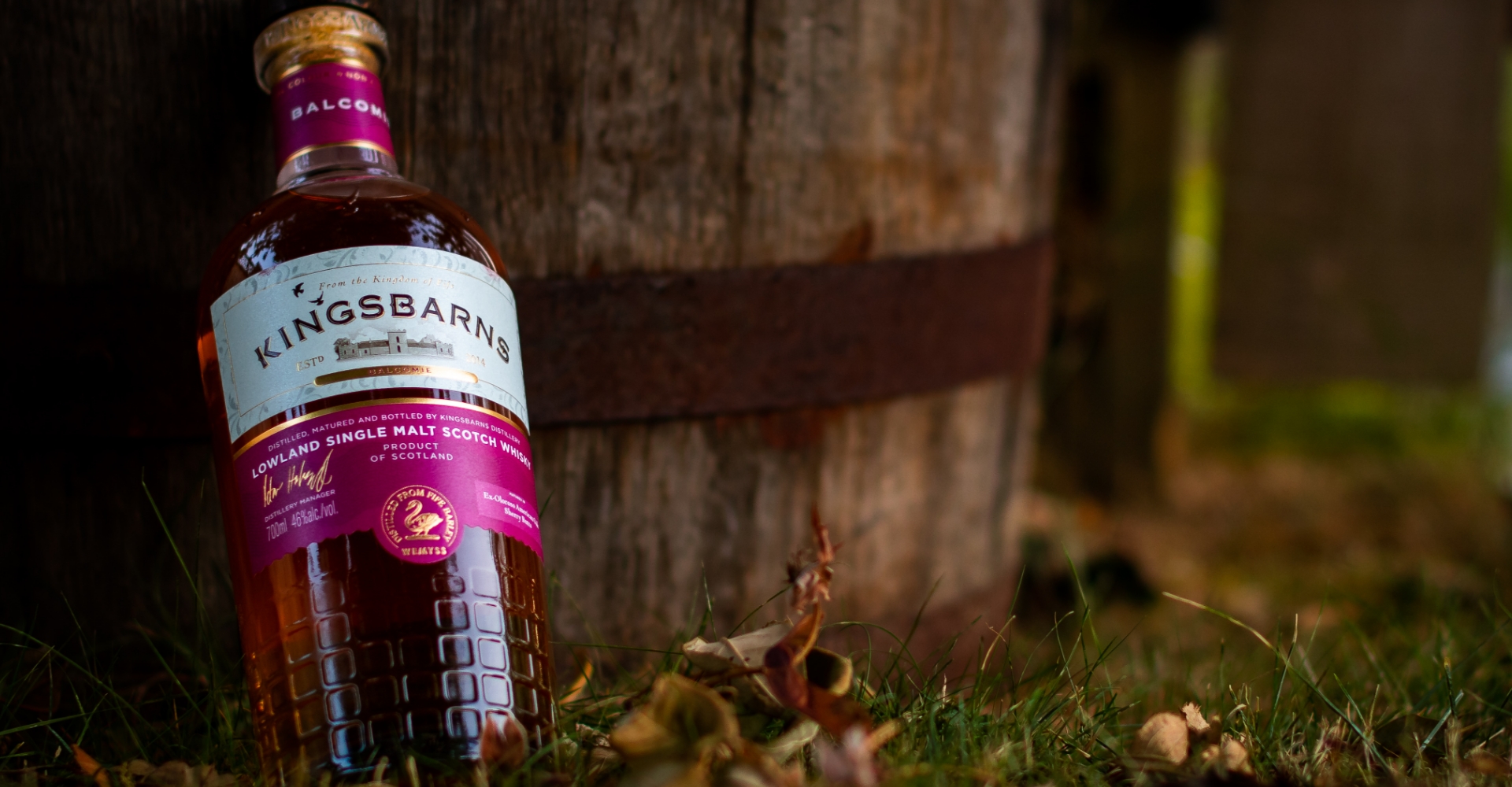 Kingsbarns Balcomie bottle with pink and gold label leaning against a brown wooden barrel with brown leaves and green grass in the foreground.