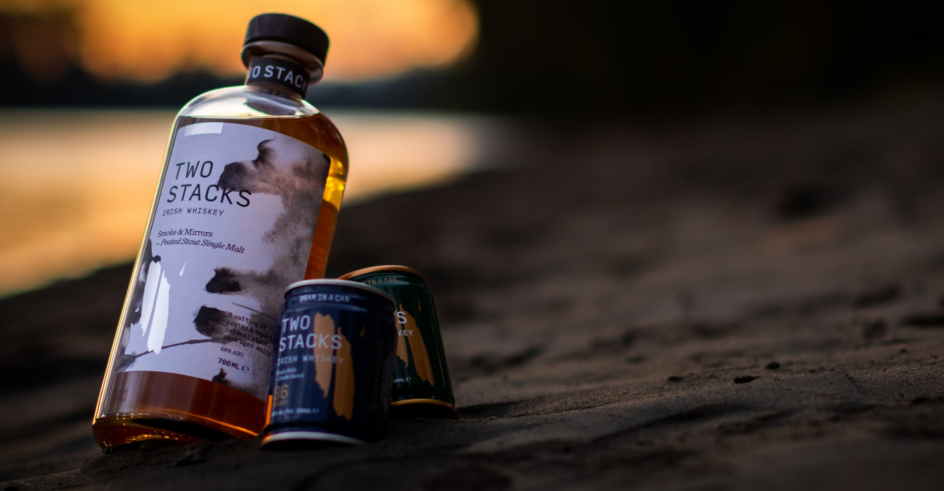 TWO STACKS15 Two Stacks Irish Whiskey bottle and blue cans sitting on sand with an orange sunset and water in the background.