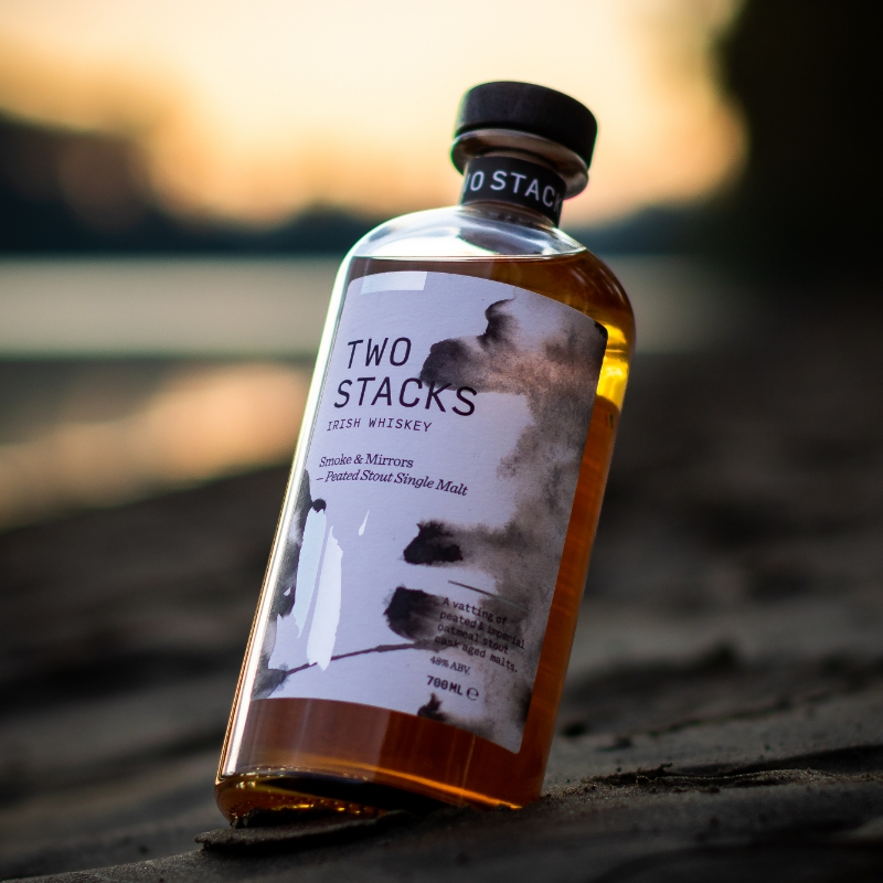 Two Stacks Irish Whiskey sitting on sand with an orange sunset and water in the background.