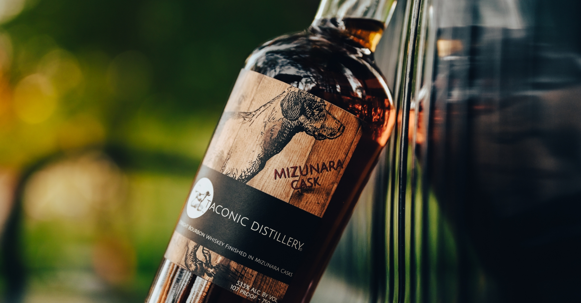 Taconic Distillery bottle leaning against a black metal bench with green foliage in the background.