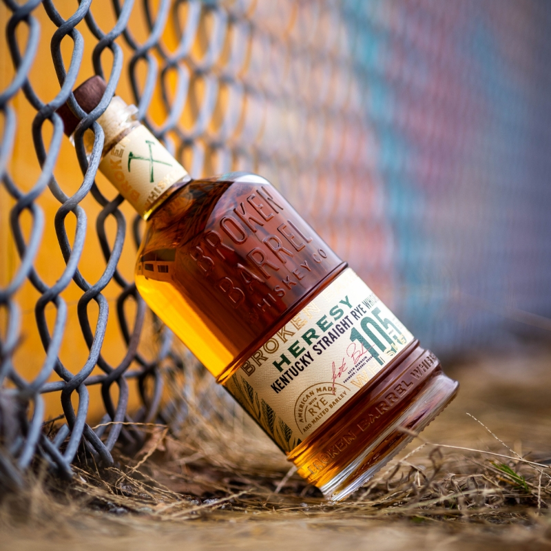 Broken Barrel Heresy Straight Rye Whiskey bottle leaning against a grey chain link fence